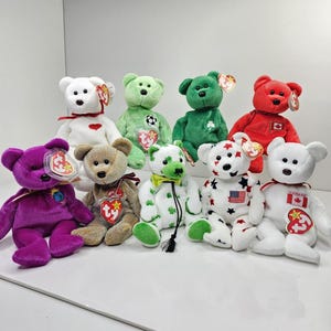 Peut inclure: Une collection de neuf ours en peluche de différentes couleurs et motifs, notamment blanc, vert, rouge, violet et beige. Chaque ours porte une étiquette rouge et jaune en forme de cœur à l'oreille, certains affichant des symboles brodés comme des drapeaux, des étoiles et des trèfles.