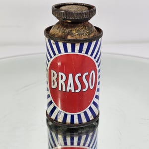 May include: Vintage Brasso metal can with a red and white label. The can features blue and white stripes and a rusty metal cap. The word "BRASSO" is prominently displayed on the front in white letters.