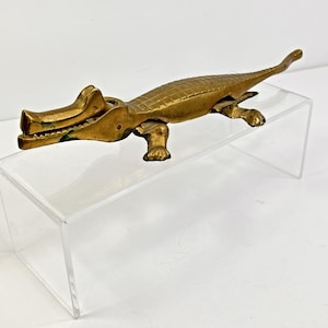 May include: A brass alligator figurine with an open mouth, detailed scales, and four legs. The figurine is displayed on a clear acrylic stand against a white background. The alligator's mouth has a green interior.