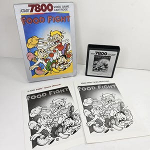 May include: Atari 7800 Food Fight video game set including a rectangular game box with colourful cartoon artwork, a black plastic game cartridge with a label, and two white paper game manuals featuring black and white illustrations of the game.