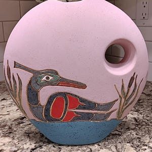 May include: A pink ceramic vase with a circular design and a large hole. The vase features a stylized bird design in blue, red, and gold, with cattails. The base is blue. The vase is signed by the artist.