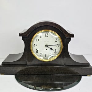May include: A dark wood Seth Thomas mantel clock with a gold-tone clock face. The clock has a white face with black numerals and hands. The clock has a curved top and a glass pendulum cover. The clock is a classic piece of home decor.