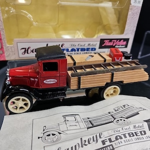 May include: A red and black die-cast metal flatbed truck model with the text "True Value" on the side. The truck has a wooden flatbed with a ladder and tool boxes. The model is 1/34 scale.