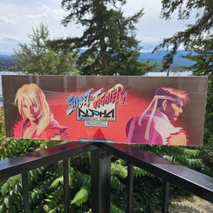 May include: A rectangular Street Fighter Alpha Warriors Dreams Capcom arcade marquee sign featuring a red background with a blonde character on the left and a brown-haired character with a white headband on the right, displaying the game title logo.