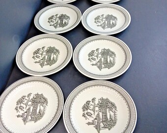 May include: A set of eight white ceramic plates with a black and white Asian-inspired landscape design. Each plate features a detailed illustration of a pagoda and trees, surrounded by a patterned border. The plates are oval and round.