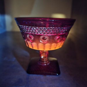 May include: A ruby red glass dessert bowl with a square base and a decorative stem. The bowl features a geometric pattern around the rim and circular designs along the sides. The glass has an orange glow from the light.
