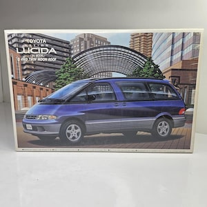 May include: A model kit box featuring a blue and silver Toyota Estima Lucida 95 Model G 4WD Twin Moon Roof van. The box art displays the vehicle parked on a paved surface in front of a modern building with a large arched structure in the background.