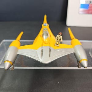 May include: A yellow and silver toy spaceship with a small pilot figure. The spaceship has a streamlined design with two silver engines and yellow wings. The pilot figure is wearing a tan uniform.