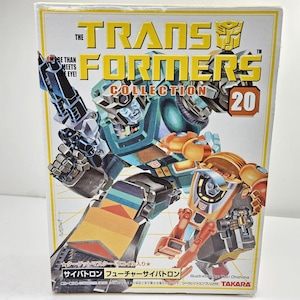 Peut inclure: Une boîte rectangulaire Transformers Collection 20 présentant une illustration de robot bleu sarcelle et noir avec une figurine de robot orange plus petite. Le texte indique The Transformers Collection 20, More Than Meets The Eye et des caractères japonais.