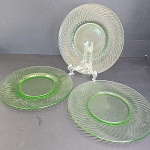 May include: Three vintage green glass plates. The plates have a spiral design around the edges and a solid green centre. One plate is displayed on a clear plastic stand. The plates are a light green colour.