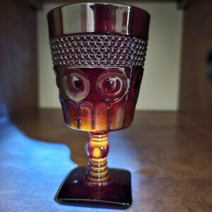 May include: A ruby-red vintage goblet with a square base and a patterned bowl. The goblet features a textured design around the rim and decorative circular patterns on the body. The stem is detailed with a series of rings.