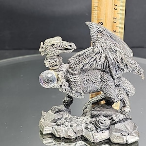 May include: A silver-colored dragon figurine with detailed scales, wings, and a crystal ball. The dragon is perched on a rocky base. The figurine measures approximately 2 inches in height.