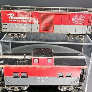 May include: Two vintage toy train cars. The top car is red, silver, and black with "Pacemaker Freight Service" text. The bottom car is red, silver, and black with "NYC 20102" text. Both cars have wheels and are displayed on a clear shelf.