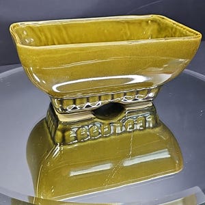 Peut inclure: Un plat en céramique vert olive de forme carrée, posé sur un socle. Le socle présente un motif décoratif à nervures. La surface brillante reflète l'environnement, créant un effet miroir.