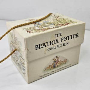 May include: A cream-coloured cardboard box featuring The Beatrix Potter Collection text and illustrations of rabbits in a garden. The box has a gold-coloured rope handle attached to the sides and includes the text The Original and Authorized Editions.