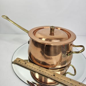 May include: A copper saucepan with a lid and a long brass handle. The pot has two small brass handles on the sides. A wooden ruler is placed in front of the pot. The pot is sitting on a mirrored surface.