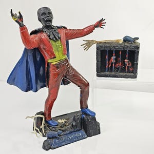 May include: A painted figurine of the Phantom of the Opera wearing a red jacket, blue cape, and green vest, standing on a dark base with a rat. Beside it is a small black stone-textured cage containing a skeletal hand and a small blue rat figure.