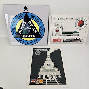 May include: Three vintage metal signs with railway themes. One sign shows the "Colorado Midland Railway" logo with a mountain scene. Another sign features the "North Coast Limited" train. The third sign depicts a steam engine.