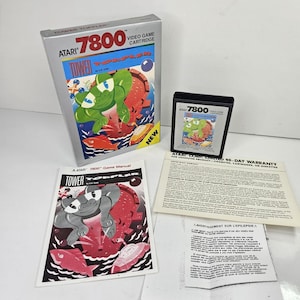 May include: Atari 7800 Tower Toppler video game set including a silver box with a green creature illustration, a black game cartridge, a game manual with a grayscale cover, and a white warranty and safety information sheet with black text.