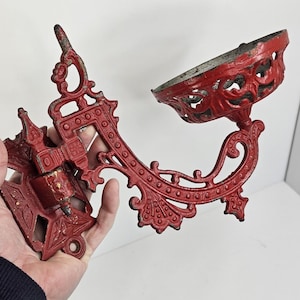May include: Ornate red cast iron wall sconce with a decorative bowl-shaped holder. The sconce features intricate scrollwork and a distressed finish, suggesting an antique or vintage style. The wall mount has a detailed design.