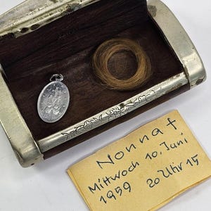 May include: An open antique wooden box with silver edging, holding a small silver pendant and a circular bundle of light brown hair. A handwritten note is present.
