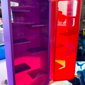 Puede incluir: Dos vitrinas, una morada y otra roja, con múltiples estantes. Las vitrinas son rectangulares con la parte superior en ángulo y están sobre una base negra. La vitrina morada tiene un interior rosa y la vitrina roja tiene un interior blanco.