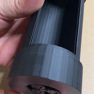 May include: A black, cylindrical container with a hollow interior. The base features a detailed, circular design. The container is held in a person's hand, showcasing its size and shape. The design appears to be 3D printed.