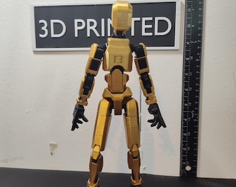 Puede incluir: Una figura de acción articulada impresa en 3D de color dorado con juntas negras. La figura está de pie sobre una superficie negra frente a una pared blanca con un letrero que dice "3D PRINTED".