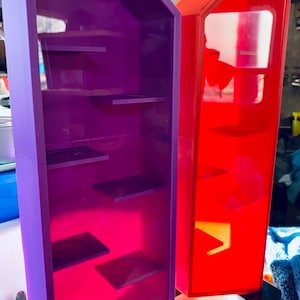 May include: Two display cases, one purple and one red, with multiple shelves. The cases are rectangular with angled tops and are set on a black base. The purple case has a pink interior, and the red case has a white interior.