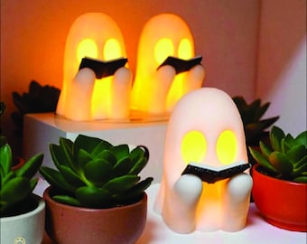 May include: Three glowing ghost-shaped night lights, each holding a small black book, are arranged on a white shelf. The lights emit a warm yellow glow. Potted succulents in various colors surround the lights.