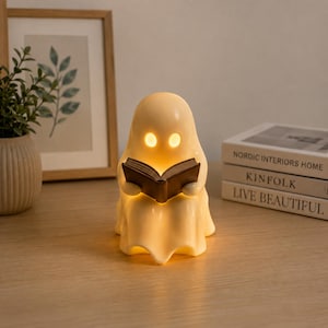 May include: A cream-colored, ghost-shaped lamp with a smooth, rounded form and two glowing oval eyes. The ghost figure is holding an open, dark brown book in its hands. The lamp emits a warm, soft light from within its hollow, translucent body.