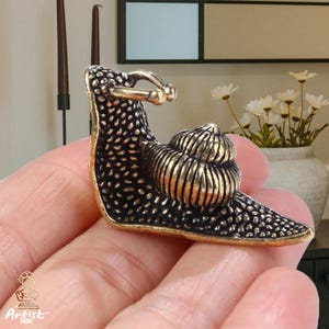 May include: A detailed bronze snail figurine with a textured body and a spiraled shell. The snail is held in a hand, showcasing its intricate design and metallic finish. The figurine has a gold-colored trim.