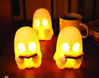 May include: Three glowing ghost-shaped night lights with open book details. The lights are yellow and have two eye-shaped openings. A yellow mug of coffee and a book are in the background. The lights are on a dark brown surface.