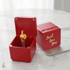 Puede incluir: Dos cajas cuadradas rojas con tapas abatibles. Una caja está abierta, revelando una figura de mano dorada sobre un resorte metálico en su interior. La otra caja está cerrada y presenta el texto dorado "Just for you" en la parte frontal.