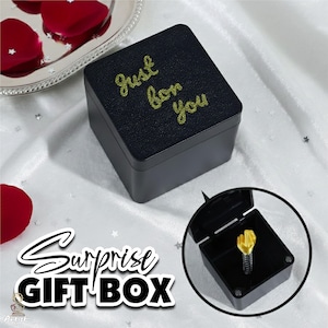 May include: Black square gift box with gold text reading "Just for You". The box is open, revealing a gold-colored object on a spring. The words "Surprise GIFT BOX" are in a stylized font.