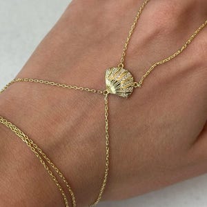 Könnte beinhalten: Ein goldenes Kettenarmband mit einem Muschel-Anhänger. Die Muschel ist mit klaren Strasssteinen besetzt.
