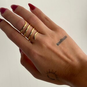 Könnte beinhalten: Ein mehrlagiger, goldfarbener Spiralring, getragen an einer Hand mit einem Tattoo, das "kindness" lautet.