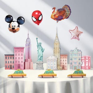 May include: A festive display featuring foil balloons shaped like Mickey Mouse, Spider-Man, a turkey, and a star. Miniature cardboard cutouts of New York City landmarks and yellow taxi cabs complete the scene, creating a celebratory atmosphere.