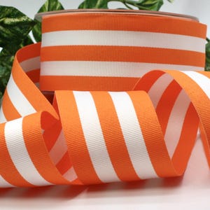 May include: A spool of orange and white striped ribbon. The ribbon features bold orange and white stripes. The ribbon is curled and displayed, suggesting its use for crafts, gift wrapping, or decorative purposes. The ribbon is likely made of a sturdy material.