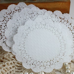 May include: A stack of white paper doilies with a delicate, lacy edge. The doilies have a central oval area with a subtle dotted texture. The background includes pearls and a crocheted surface, suggesting a vintage or craft theme.