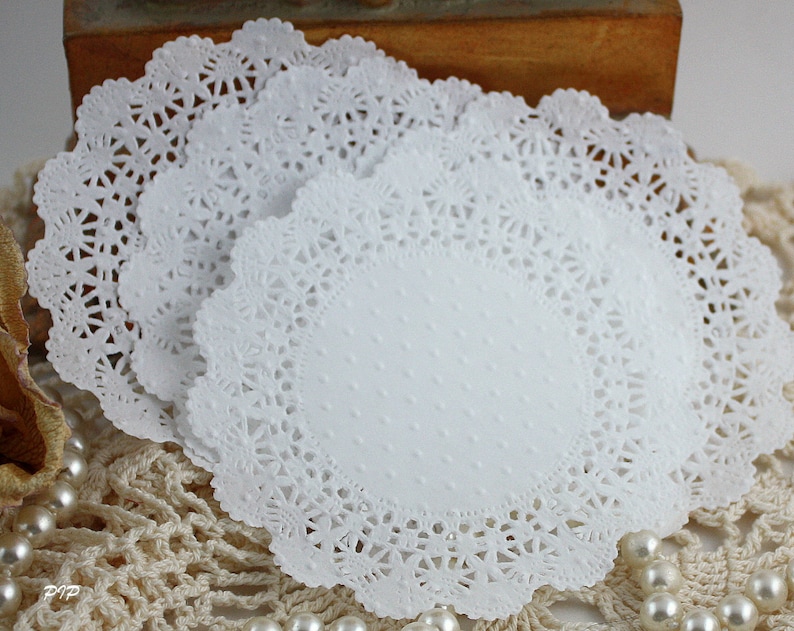 May include: A stack of white paper doilies with a delicate, lacy edge. The doilies have a central oval area with a subtle dotted texture. The background includes pearls and a crocheted surface, suggesting a vintage or craft theme.