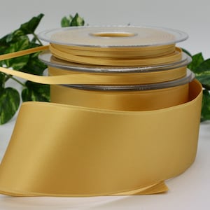 May include: Several spools of gold satin ribbon. The largest spool is unrolled, showing the smooth texture and sheen of the ribbon. The ribbon is a warm, golden colour, ideal for crafts and gift wrapping. The ribbon widths vary.