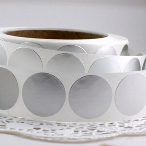 May include: A roll of silver metallic circle stickers on a white backing. The stickers are partially unrolled, revealing the shiny, reflective surface. The roll sits on a white lace doily.