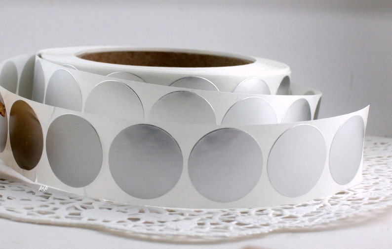 May include: A roll of silver metallic circle stickers on a white backing. The stickers are partially unrolled, revealing the shiny, reflective surface. The roll sits on a white lace doily.