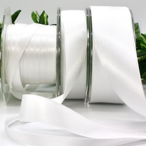 May include: Three spools of white satin ribbon. The ribbon is unwound from the spools, with the ends of the ribbon lying on a white surface. Green leaves are visible in the background.
