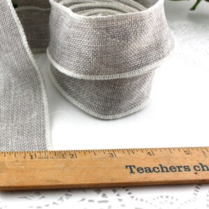 May include: A roll of gray ribbon with white edging, next to a wooden ruler. The ribbon is made of a textured fabric. The ruler is marked in inches and has the text "Teachers change lives."