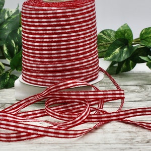 May include: A spool of red and white gingham ribbon. The ribbon is displayed on a white surface with green leafy plants in the background. A length of the ribbon is unrolled and lies in front of the spool.