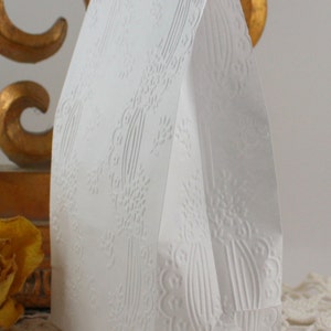 May include: A white paper gift bag with an embossed floral design. The bag has a folded top and is standing upright. The embossed pattern includes floral and vertical line elements, creating a textured surface.
