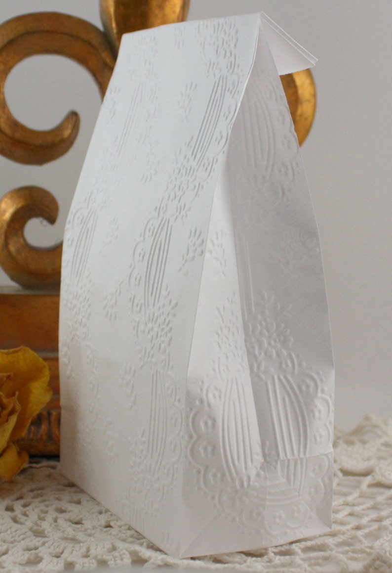 May include: A white paper gift bag with an embossed floral design. The bag has a folded top and is standing upright. The embossed pattern includes floral and vertical line elements, creating a textured surface.