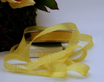 May include: A spool of yellow ribbon with a textured, semi-transparent appearance. The ribbon is unrolled and draped across a white surface, with a roll of ribbon in the background. A yellow rose and greenery are in the background.